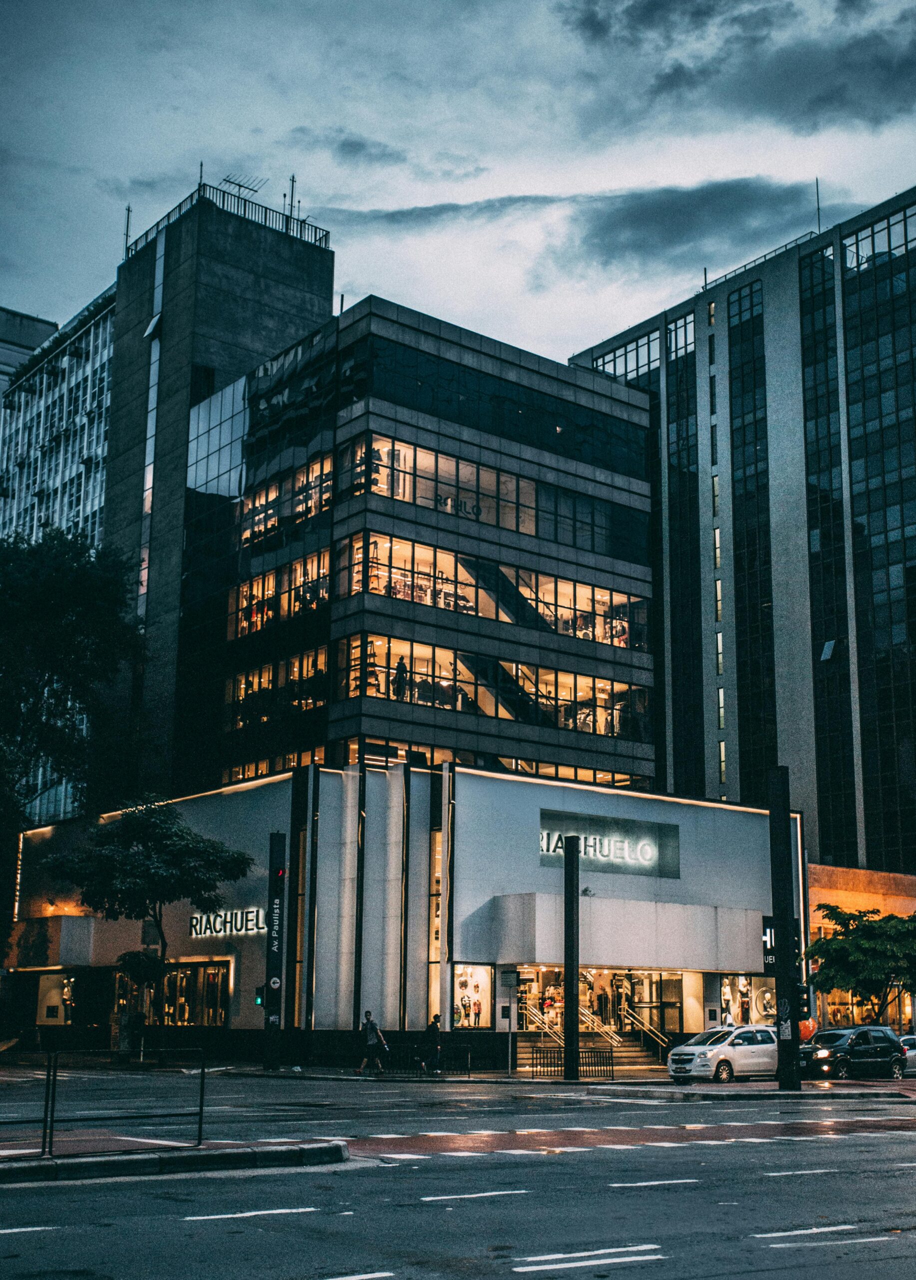 Night view of a city block with illuminated store, modern architecture, and cloudy sky.